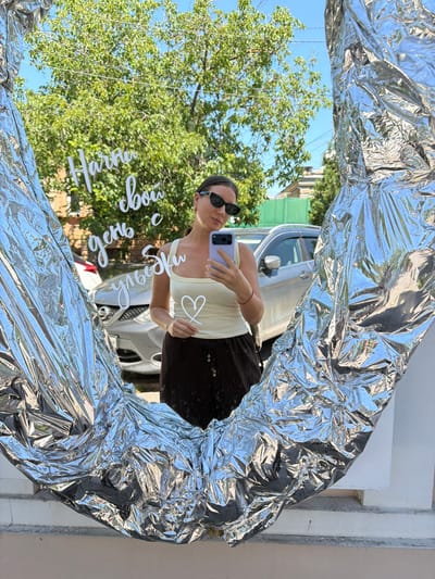 A woman is taking a selfie in a mirror that is framed by crinkled silver material. The woman is wearing sunglasses and a light-colored tank top, and she is holding up her phone to take the picture. In the reflection of the mirror, we can see a car parked behind her, as well as some trees and a blue sky. There is white text written on the mirror that appears to be in Russian. The text reads "Начни свой день с улыбки" which translates to "Start your day with a smile." The woman is smiling in the picture. The overall scene appears to be outdoors, possibly on a sunny day. The location is Taganrog, Russia.