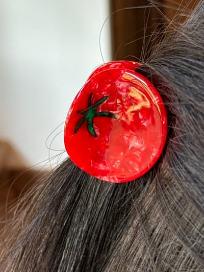 A close-up image captures a section of dark, fine hair, possibly black or dark brown, with individual strands visible. A circular, glossy red hair accessory, approximately 2-3 cm in diameter, is affixed to the hair on the right side of the frame. The accessory features an opaque green, star-shaped design on its surface, mimicking a tomato's calyx. The red material exhibits a marbled or textured pattern beneath its clear, reflective top layer. The background is uniformly light and out of focus on the left, transitioning to a darker, indistinct blurred area on the right, suggesting an indoor environment. No specific features within the image indicate a precise location in the city of Otego, United States.