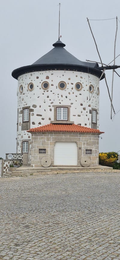 The image depicts a traditional stone and white-plastered windmill in Póvoa de Varzim, Portugal. The windmill has a conical, dark gray roof and its cylindrical body is adorned with round, small windows and irregularly placed stones. A portion of the windmill's blades are visible on the right side of the frame. At the base of the windmill, there is a section with a red-tiled roof supported by a stone structure. This section features a large, white roller door and two large millstones positioned on either side. The ground in front of the structure is paved with cobblestones. The sky is overcast.