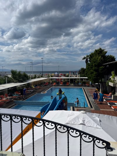 The image depicts a resort pool area in Ayvalık, Turkey, on a cloudy day. In the foreground, a black wrought-iron railing partially obscures a white umbrella. Beyond the railing, the resort's blue swimming pool is visible, with a blue water slide. Several people are in and around the pool, including children playing on the slide and in the water. Lounge chairs and umbrellas are set up around the pool and on the surrounding tiled patio. In the background, palm trees and lush green trees are present. Further back, there is a view of the beach and the sea. The sky is filled with dramatic clouds. A hotel sign reading "Hava Hotel" is visible in the distance.