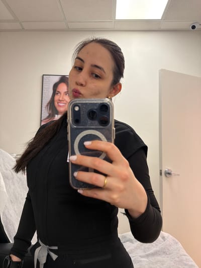 A medium-shot, eye-level, indoor photograph depicts a woman taking a selfie in a room that appears to be a medical or aesthetic treatment room.
The woman is holding a smartphone with a clear case and a magnetic charging ring. She is wearing a black uniform with a long-sleeved top and pants. Her hair is pulled back, and she has a gold ring on her finger.
Behind the woman, a poster on the wall advertises "Nuskin". A medical or aesthetic treatment bed covered with a white sheet is also visible. A security camera is mounted on the wall near the ceiling. A silver-colored door handle is visible on a white door.
Given the "Nuskin" poster and the treatment bed, the location appears to be a medical spa or aesthetic clinic in an unknown location in the city of Las Vegas, United States of America.