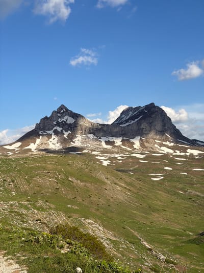 The image shows a mountain in the city of Grabovica, Montenegro. The mountain has a rugged, rocky face with patches of snow clinging to its sides. The lower slopes are covered in green vegetation. The sky is a bright blue with scattered white clouds.