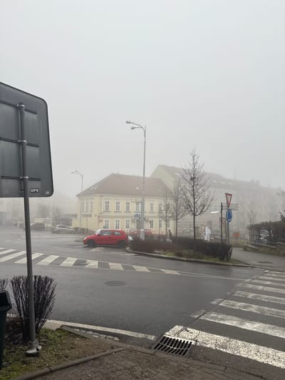 The image depicts a foggy street intersection in Znojmo, Czechia, under an overcast sky. The overall visibility is reduced by a dense, grey fog. The asphalt road surface appears wet, reflecting ambient light.

In the immediate foreground on the left, the rear of a dark, rectangular signpost is visible, affixed to a metallic pole, with "UFX" text printed on its lower left. A small, dark green bush grows at the base of the pole, and a green trash receptacle is partially visible to its left. On the right foreground, a metal storm drain is set into the concrete sidewalk.

The midground features a pedestrian crossing marked by white "zebra" stripes across the wet asphalt. A bright red two-door hatchback car, appearing to be an older model, is positioned at the intersection's corner, facing right, seemingly stopped or parked. Adjacent to the road, a raised, landscaped median contains several leafless deciduous trees and low, dark green shrubs. A tall, grey streetlamp with two light fixtures stands prominently in the center-right of the frame; another, smaller streetlamp is visible further down the street on the left. Multiple traffic signs are present on poles, including a red triangular yield sign and a blue circular sign.

In the background, a two-story yellow building with a reddish-brown tiled roof and numerous white-framed windows is prominent, suggesting a commercial establishment with faint text on its facade. Behind and to the right of this building, a taller, multi-story residential building with a darker facade and multiple windows is partially obscured by the fog. Several other vehicles, mostly grey or silver, are parked along the street further to the left. No individuals are visible in the scene.