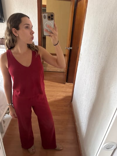 A female subject is captured in a mirror selfie within an interior residential setting in Ciudad Victoria, Mexico. The exact location within the city is not discernible from the image. The subject, a Caucasian female with light brown, wavy hair, appears to be in her mid-20s to early 30s. She is dressed in a matching deep red sleeveless V-neck tank top and wide-leg pants, complemented by light-colored open-toed sandals. She wears small white stud earrings, a thin necklace, and a silver-toned watch or bracelet on her left wrist. The subject holds a light-colored smartphone, consistent with an iPhone 15 Pro Max due to the visible camera array and MagSafe ring on the case, in her right hand to capture the reflection. She is depicted with her head slightly tilted, looking at her reflection, and her lips are puckered. The background visible in the mirror includes a polished light-brown wooden floor, a white textured wall on the right, and a dark wooden door with a silver knob. A portion of a white bed frame or furniture is visible on the far left. The lighting is even and bright, indicating an illuminated indoor space.