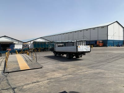 The image depicts an industrial or commercial facility in Babawa, Nigeria, under a clear daylight sky. The scene is dominated by a large, paved asphalt or concrete lot showing visible tire tracks.
In the foreground, to the left, is a dark-colored vehicle scale or ramp with yellow anti-slip stripes in the center and yellow guardrails on both sides, inclined upwards from left to right. Immediately to its right, a light-colored flatbed truck with an open, dark-colored cage bed is positioned, facing away from the viewer. The rear of the truck displays some wear and minor damage.
In the mid-ground, to the left, a large, light-colored truck with a blue-green tarp or container on its bed is parked near a building with a light-colored roof and blue accents. Further back, on the right side of the frame, is a long, single-story warehouse building with white walls and a distinct dark blue band along its lower section. Its roof is light-colored.
Parked in front of this main warehouse building, further back in the mid-ground, is an orange truck. To the far right, two blue passenger cars are parked against the warehouse wall. One individual is visible standing near the orange truck. Several yellow bollards and tall lamp posts are distributed across the paved area.