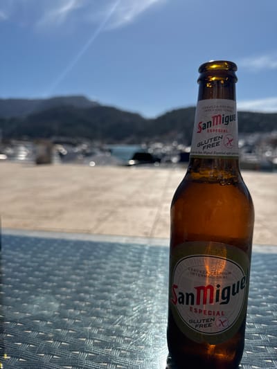A brown glass bottle of San Miguel Especial Gluten Free beer, exhibiting condensation, stands prominently in the right foreground on a light blue woven table surface. The scene is located at a marina or port area within El Port, Spain. The midground depicts a light-colored paved waterfront, extending towards a body of water in the background. Numerous white boats and yachts are docked in the water, appearing out of focus. Beyond the marina, a range of dark, forested mountains is visible under a clear, bright blue sky with sparse, wispy clouds and a faint contrail. The lighting suggests full daytime. No individuals are present in the image.
