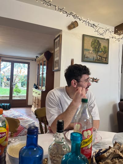 The image depicts an indoor domestic scene set within a residence in Ancenis, France. A single male individual is seated at a table in the mid-ground, observed from the chest up. He wears a white t-shirt, has dark, curly hair, eyeglasses, and a mustache, and is looking towards the right side of the frame with his right hand near his chin.
The table in the foreground is covered with a white tablecloth and features several items. These include a partially empty Schweppes Pomme (apple) bottle with a red label, a Lipton Ice Tea (peach) bottle, and multiple empty or near-empty glass bottles of varying colors (blue, clear, and one labeled "Kraken"). A white bowl containing a light-colored substance and a basket filled with sliced bread are also present on the table.
The background reveals white walls decorated with string lights draped across the ceiling and a wall. A large, wooden hutch with glass cabinet doors and drawers stands to the left, displaying dark figurines, possibly ducks, on a shelf, and a purple decorative lantern hanging from it. A dark-framed picture is mounted above the hutch. To the right of the hutch, a framed painting depicting a landscape with trees, water, and huts hangs on the wall, below which a decorative wooden fork-like object is affixed. A small, patterned, cylindrical vessel is also mounted on the wall near the ceiling. A segment of a brown leather sofa or chair is visible on the far right. The floor consists of wooden planks partially covered by a patterned rug that extends towards a set of double glass doors.
Beyond the glass doors, an outdoor garden is visible. It features green grass, trees with sparse foliage, a sculpted evergreen shrub, and a low stone wall. A blue and black rolled mat or bag lies on the floor near the base of the glass doors.
