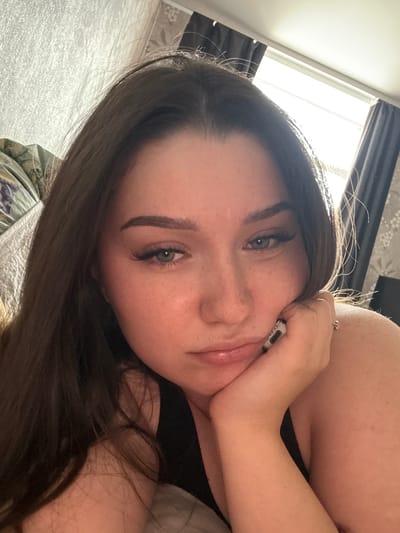 A young woman with long, dark brown hair is resting her head on her hand, looking down with a pensive expression. Her skin is fair with a slight blush on her cheeks. She has noticeable makeup, including winged eyeliner and false eyelashes. A ring is visible on her right ring finger. She is wearing a black top. The background shows a textured wall and curtains drawn across a window. The scene appears to be indoors, likely in a bedroom, possibly in the city of Гамово, Russia, judging by the provided context.