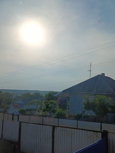 The image depicts a rural or suburban scene in Podvirne, Ukraine, under a bright, overcast sky. The sun is visible as a large, diffused bright spot in the upper left quadrant, suggesting it is daytime, likely morning or late afternoon due to the soft light. The sky is a pale blue with wispy clouds.
The foreground is dominated by a corrugated metal fence, with a section of a more decorative fence with ironwork visible behind it. Beyond the fence, a residential area unfolds. Several houses with dark, sloped roofs are visible, with one house in the mid-ground having a prominent television antenna on its roof. Lush green trees and some flowering bushes, including a large bush with purple flowers, dot the landscape around the houses, indicating a spring or summer season. In the distance, a gentle, rolling landscape of fields and trees stretches to the horizon.
There are no people or specific activities clearly depicted. The scene is serene and quiet, conveying a sense of everyday life in a village setting. The overall mood is calm and peaceful, bathed in soft, natural light. No visible text is present in the image.