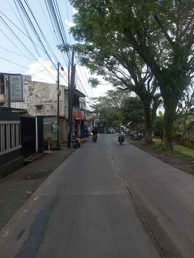The image shows a straight, asphalt road leading into the distance, flanked by buildings on the left and trees on the right. Two motorcycles are traveling on the road. Power lines crisscross overhead. A sign for "Jehan Beauty & Style" is visible on a building to the left. The road surface appears somewhat worn with a central crack. Other parked motorbikes are visible further down the road. The buildings on the left appear to be in a mixed state of repair. The trees on the right are tall and overgrown, obscuring parts of the landscape. The image does not provide any information to identify the location within Xayabury District, Laos.