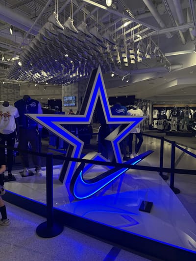 A retail store displays Dallas Cowboys memorabilia. A large star logo, lit with blue neon, takes center stage, accompanied by a blue neon Nike swoosh on a nearby display. Racks of clothing and sports jerseys are visible in the background, alongside a large arrangement of white sneakers suspended from the ceiling. The lighting creates a bright and modern retail environment.