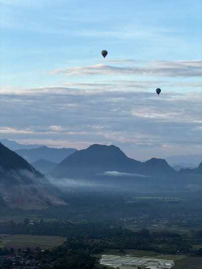 The image captures a serene aerial landscape of Naka, Laos, during what appears to be early morning. Two hot air balloons, one with blue, yellow, and red stripes and another with blue and yellow stripes, are visible in the soft blue sky, floating above a layer of hazy clouds. The landscape below is dominated by lush green mountains and valleys shrouded in a light mist. In the foreground, a village with houses and rice paddies can be seen. The sun is not directly visible, suggesting it is either before sunrise or just after, with the sky illuminated by diffused light. The overall scene is tranquil and picturesque, showcasing the natural beauty of the region.