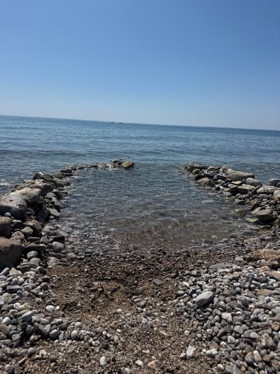 The image shows a pebble beach and a calm body of water under a clear blue sky. Two small boats are visible in the distance on the water. The foreground is filled with various sizes of pebbles and sand, with a break in the pebble line leading into the water, forming a small inlet. The water is clear, allowing the seabed of pebbles to be visible. The scene is tranquil and depicts a natural coastal environment. The location is specified as the city of Đurovići, Montenegro.