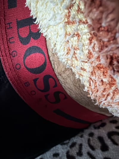 A close-up, angled shot shows a red elastic waistband with the word "BOSS" in large black lettering. The fabric appears to be textured, possibly cotton. A fuzzy, cream-colored material with reddish-brown speckles is draped over the waistband, obscuring part of it. A patch of dark hair is visible beneath the fuzzy material, adjacent to the waistband. The background is dark, with a glimpse of a leopard-print pattern at the bottom right. The image does not provide any information about a location in Avdarma, Moldova.