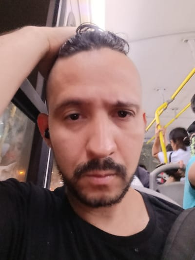 A selfie depicts a man on public transport, possibly a bus. He wears a black t-shirt and sports a mustache and neatly trimmed beard. His dark hair is styled in a short mohawk. His hand rests on his head. Other passengers are visible in the background, and a yellow handrail is in the periphery. The overall image has a casual, slightly weary tone.