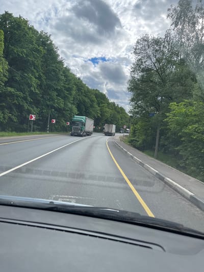 The image depicts a road in Semyonovskoye, Russia, viewed from inside a vehicle. The road has multiple lanes marked by white and yellow lines. Several trucks are traveling in the same direction. The roadside is lined with dense green trees and red-and-white directional signs. The sky is visible with scattered clouds. A portion of the car's interior, including the windshield wiper, is visible in the foreground.