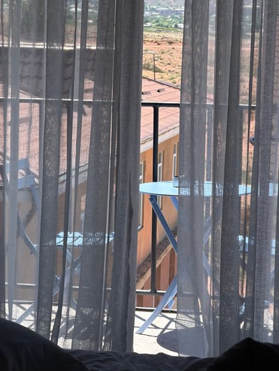 This is a close-up shot of sheer grey curtains obscuring a view of an outdoor balcony area in Kaji-Say, Kyrgyzstan. Through the translucent fabric, a light blue metal table with a similar blue metal chair are visible. The table has a small silver object on it. Beyond the balcony, there is an orange building with white-framed windows and a tiled roof. In the background, a distant landscape shows arid, tan-colored hills and some scattered vegetation under a bright sky. A black railing runs horizontally across the middle of the frame. The bottom of the image shows a dark, out-of-focus foreground of what appears to be bedding.