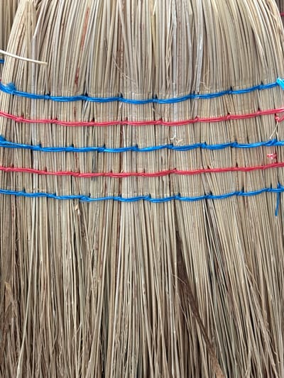 This media file presents a close-up, overhead view of a bound bundle of dry, straw-like material, likely broomcorn or a similar dried grass. The material is tightly packed and arranged horizontally, with individual strands clearly visible. Running vertically across the bundle are several strands of brightly colored twine, alternating between red and blue. These strings are knotted at intervals, securing the bundle and creating a textured pattern. The overall color palette is muted, dominated by the beige and tan tones of the dried material, with pops of red and blue from the twine. There are no people, specific activities, or discernible location cues beyond the material itself. The lighting appears even, suggesting it was taken in daylight, but no specific time of day or weather conditions can be determined. No text is visible in the image. The image focuses entirely on the texture and construction of this bundled material.