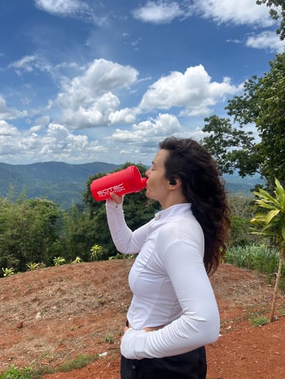 A woman with long, curly dark hair is shown from the mid-torso up, in profile, drinking from a red shaker bottle. She is wearing a white long-sleeved shirt and black pants, and black earbuds are visible in her ears. The background shows a lush, mountainous landscape under a bright blue sky with scattered white clouds. The woman is standing on a reddish-brown dirt slope, with green trees and foliage around her. The location is stated to be Nao Sẻ, Vietnam.