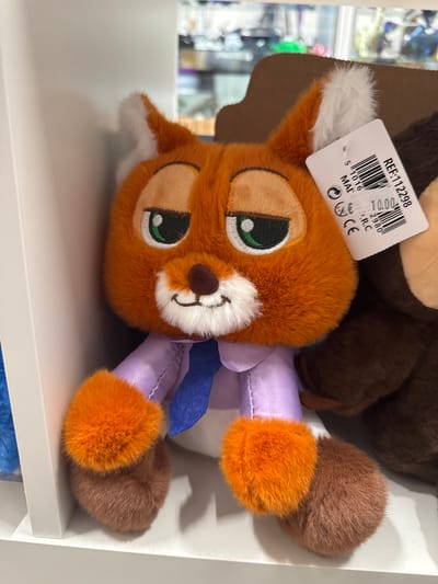 The image shows a close-up of a plush toy, possibly a fox, lying on a white shelf. The toy is predominantly orange with green eyes and a white muzzle. It is wearing a light purple outfit with a blue sash. The toy is lying on its side, with its head and body angled towards the camera. Behind the fox, a portion of another plush toy, appearing to be brown and larger, is visible. A tag is attached to the fox's ear.

The tag has a barcode and text printed on it. Visible text includes "REF:112298", "70.00", "5 1016", "MAD", "IN", ".R.C", "CE", and a symbol that resembles the Euro currency.

The background is out of focus but appears to be a retail setting, with shelves filled with other items. The lighting suggests an indoor environment, likely during daylight hours given the brightness of the scene. The overall impression is one of a product display in a store. The location context provided is Keila, Estonia. There are no people visible in the image. The toy is stationary, with no activity taking place.