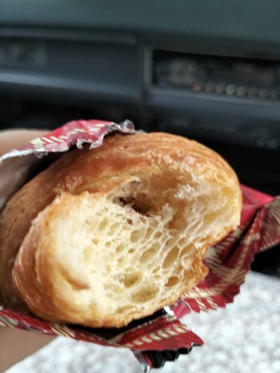 A partially consumed, light brown, croissant-like pastry is depicted in close-up, held by an unseen individual's hand in Gleputoh, Indonesia. The pastry features a bite taken from its left side, exposing an airy, irregular crumb structure typical of laminated dough, with a dark brown, possibly chocolate, filling visible within. It is partially unwrapped from a crinkled red foil wrapper adorned with a gold pattern. The blurry background indicates the interior of a vehicle, with a dark, indistinct dashboard or control panel at the top and a lighter, textured surface below.