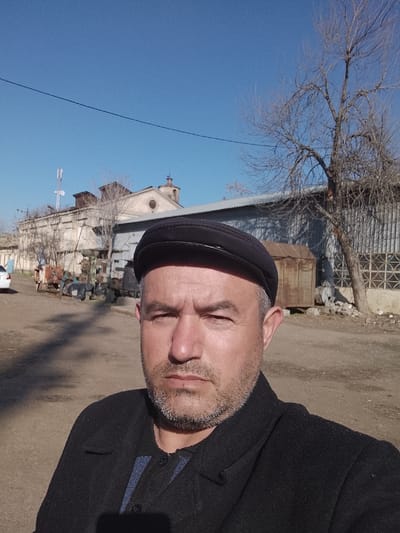 A man is centered in a selfie taken outdoors in Baxt, Uzbekistan, under a clear blue sky. He is approximately 40-50 years old, with light facial stubble, wearing a black flat cap, a black coat, and a dark blue striped shirt. He looks directly at the camera with a neutral expression. The background features an unpaved dirt area. To the left, a partial white vehicle is visible near a collection of metal objects or machinery parts. Further back, a light-colored, aged building with a small cupola and a white cell tower on its roof stands next to a corrugated metal building. Several bare trees are present, including one prominent tree on the right side. A weathered metal container sits near the corrugated structure. A power line traverses the upper frame.