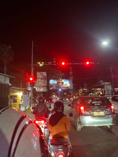 The image captures a nighttime scene on a street in Macege, Indonesia, with heavy traffic congestion. The foreground is dominated by the rear of a motorcycle, its rider visible in a black helmet and an orange jacket. To the right, another motorcycle with a rider wearing a black helmet and hijab is also stopped. A silver Toyota car, with the license plate DW 1467 EF, is in front of the second motorcycle. The street is illuminated by red traffic lights, street lamps, and the headlights of vehicles. In the background, several buildings and billboards are visible, including a mosque with a minaret. The atmosphere is dark, with a red glow emanating from the traffic signals and ambient lighting from the city.
