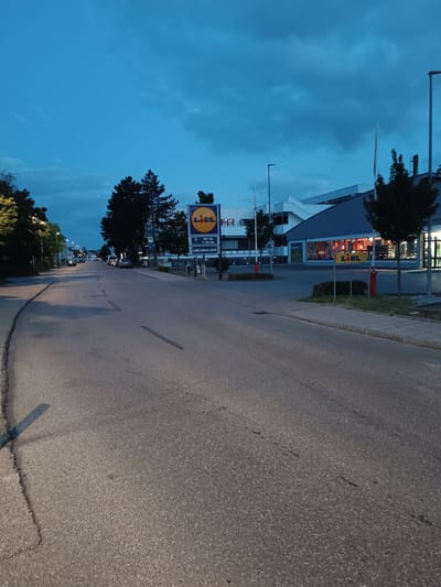 The image depicts a street scene in Germering, Germany, likely at dusk or early evening due to the dim lighting and the blue hue of the sky, which has some scattered clouds. The street is asphalt and appears to be a two-lane road with a sidewalk on the right side.
On the right side of the street, a Lidl supermarket is visible. It has a modern architectural design with a dark grey roof and large windows. Several trees are planted in front of the building. Two Lidl signs are visible: one larger sign on a pole with the store's logo and opening hours (Mo-Sa 7-20 Uhr), and a smaller yellow sign with the Lidl logo closer to the road. There are also red bollards and a small hedge along the sidewalk.
On the left side of the street, trees and some residential buildings are visible. Further down the street, there are parked cars and streetlights. A few vehicles are driving on the road in the distance. The overall environment suggests a typical suburban or commercial area of a German city. There are no people clearly visible in the image, and no discernible actions or interactions are taking place. The scene is static and appears to be captured from a slightly elevated perspective, looking down the street.