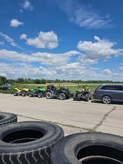 The image depicts a scene on an outdoor paved area, likely a parking lot or track, with a row of ATVs parked in the middle ground. In the foreground, large tires are stacked. Behind the ATVs, a grassy field stretches out, bordered by trees in the distance. A dark gray station wagon is parked to the right of the ATVs. The sky above is bright blue with scattered white clouds. The location is identified as Akatsievo, Bulgaria.