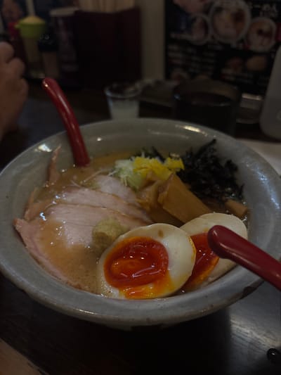 This image presents a close-up, slightly elevated view of a bowl of ramen in an indoor restaurant environment. The ramen is contained within a light grey, textured ceramic bowl. The broth is light brown and opaque, with visible oil droplets. Toppings include multiple thin, light pink slices of pork chashu, two halves of a soft-boiled ajitama egg with bright orange, semi-liquid yolks, and firm white albumen. Other visible toppings are elongated pieces of light brown menma (bamboo shoots), dark, thin strips consistent with kikurage mushrooms or seaweed, a small mound of finely minced green onions, a cluster of bright yellow shredded material, and a small dollop of light green paste situated on a pork slice. A red-handled spoon is positioned inside the bowl, pointing towards the upper left. A second red-handled spoon is partially visible in the lower right, outside the bowl. The background is blurred, showing a portion of a person's hand on the left, various table condiments including a green shaker and a dark caddy in the center-left, and a menu board featuring Japanese text and illustrations of ramen bowls in the upper right. The scene is illuminated by artificial interior lighting. While the image is consistent with a ramen restaurant in Japan, no specific visual identifiers are present to forensically determine the exact location within the city of Kanazawa.