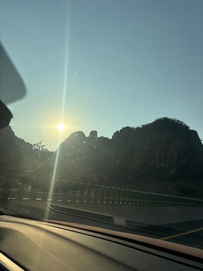 The image captures a view from inside a vehicle on a highway near Tepoztlán, Mexico, during either sunrise or sunset. The sun is a prominent, bright orb with a strong sunburst and vertical lens flare, positioned low in the left-center of a clear, light blue sky. Dark, rugged mountains, characteristic of the Sierra de Tepoztlán, are silhouetted against the bright sky in the mid-ground and background, showing some sparse vegetation. In the lower mid-ground, a multi-lane road or highway is visible, separated by a concrete median or barrier. A green-panelled sound barrier or fence runs parallel to the road on the right. The vehicle's dashboard is visible across the bottom of the frame, and a partial view of a sun visor or similar interior object is present in the upper left corner. No people or other vehicles are explicitly visible in the external environment. Scattered light particles, possibly dust on the windshield or lens artifacts, are present throughout the sunlit areas.