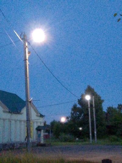 The image captures an outdoor scene at dusk, likely in Savino, Russia, given the provided context. A prominent wooden utility pole with a lit street lamp stands in the foreground, slightly to the left of center. Power lines extend from the pole across the sky. To the left of the pole, a portion of a building with a dark green roof and light-colored walls is visible. In the background, several trees are silhouetted against the deep blue evening sky, with another street lamp illuminated among them. A faint, circular light source, possibly the moon, is visible in the upper right portion of the image. The ground in the foreground appears to be a mix of vegetation and a paved surface.