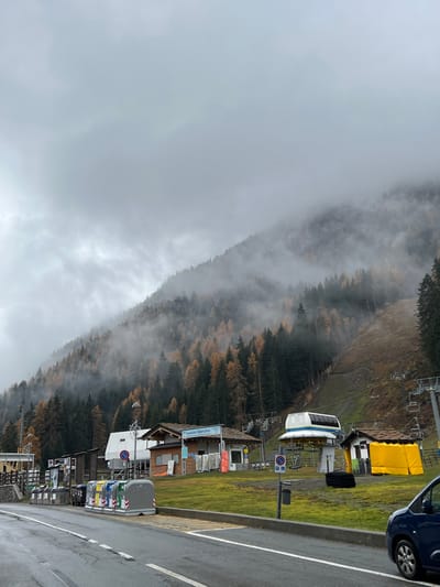 An outdoor scene in La Thuile, Italy, depicts a foggy, overcast mountain environment. In the foreground, a wet asphalt road runs horizontally with a double white centerline and a single white shoulder line. A curb separates the road from a grassy area where a row of multi-colored recycling bins (green, yellow, blue, grey) and a grey parking sign pole are present. The front right section of a dark blue compact car is visible on the far right of the road.

The midground features a sloping grassy expanse. Several buildings are positioned here: a small wooden building on the left, a larger wooden structure with a gabled roof and a sign reading "Maison Blanche" in the center-left, and another small building with a dark roof connected to a yellow tent-like structure on the right. A prominent white and blue ski lift station, possibly a gondola or chairlift terminal, is situated on the grass to the right of the "Maison Blanche" building. Multiple ski lift towers and empty chairs extend upwards from this area into the mountain.

The background is dominated by a densely forested mountain slope. The forest consists of dark green evergreen trees interspersed with numerous deciduous trees displaying vibrant orange and yellow autumn foliage. Heavy fog and low-hanging clouds partially obscure the upper sections of the mountain and the forest, creating a muted, atmospheric effect. A cleared ski slope, appearing as a lighter brown-green strip, descends on the right side of the mountain through the trees and fog. No individuals are visible.