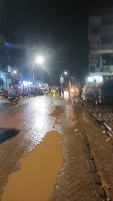 This image depicts a nighttime street scene within Kisii Central ward, Kenya. The paved road, constructed of bricks or cobblestones, is thoroughly wet, featuring numerous large puddles that reflect ambient light sources. A bright overhead streetlight dominates the central mid-ground, illuminating the wet surface.
On the left side of the street, multiple motorcycles are parked along the curb, with one motorcycle and rider visible in the foreground. Commercial buildings line this side, displaying illuminated signs, including a partially legible "RALPH'S" sign. An elevated metal structure, possibly a pedestrian bridge or ramp, is also visible extending from the left.
On the right side, more multi-story commercial buildings are present, along with a parked vehicle, possibly a truck or van. Piles of what appear to be wooden planks or debris are stacked along the roadside.
Several pedestrians are visible walking on the street in the mid-ground, moving in various directions. A distant vehicle's headlight is visible further down the road. The scene is illuminated by streetlights, vehicle lights, and lights emanating from the commercial establishments, all reflecting off the water-covered road surface. The conditions suggest recent rainfall.
