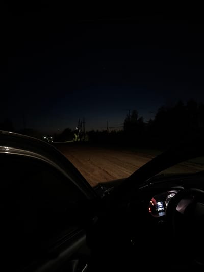 The image, taken at night in what appears to be Borodulino, Russia, shows a view from inside a vehicle. The vehicle's door is open, revealing a dirt road illuminated by a few distant streetlights and building lights. The dashboard is visible, with illuminated indicators and displays. The background consists of a dark sky, trees, and utility poles along the roadside.