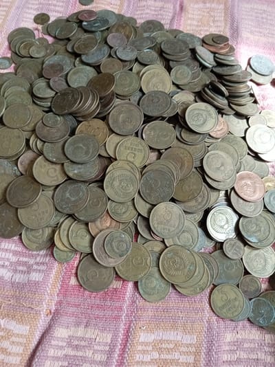 The image shows a large pile of old, tarnished coins, primarily silver and copper colored, scattered across a textured pink and white fabric. Many of the coins are worn and some are stacked. The coins appear to be of Soviet origin, with visible markings such as the hammer and sickle emblem and Cyrillic script. There are no people or animals present in the image, and the environment is limited to the surface on which the coins are placed. The image does not contain any information that would allow for the identification of a specific location in Bugulma, Russia.