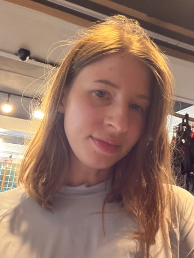 The image shows a young woman with medium-length, light brown hair, taken from a close-up perspective. She is wearing a light grey long-sleeved shirt. The background is blurred but appears to be indoors, with ceiling lights and some racks of dark items visible on the right side. There is no discernible information about the location being in Seo-myeon, South Korea.