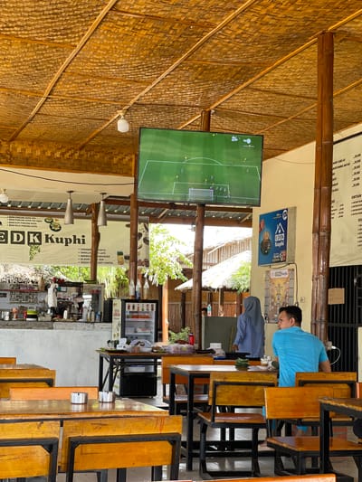 The image depicts the interior of an open-air restaurant with a thatched roof, located in Julikeude Dua, Indonesia. The atmosphere is casual, with wooden tables and chairs arranged for diners. A large television screen mounted on the wall displays a soccer match, suggesting a place where people gather to watch sports.
In the foreground, two individuals are seated at a table, engrossed in their own activities. A man in a blue shirt is looking away from the camera, while another person, partially visible, is facing the television. In the background, a woman in a light blue hijab is standing near a counter area, possibly attending to customers or preparing food.
The restaurant is equipped with a refrigerator displaying various beverages, and a counter stocked with utensils and condiments. The overall impression is that of a local eatery offering a relaxed dining experience, possibly catering to both locals and tourists. The presence of a menu board with prices in Indonesian currency further reinforces the authenticity of the establishment.
