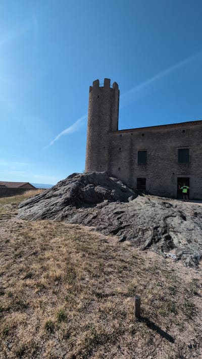 The image depicts the medieval castle of Chambles, France, under a clear blue sky. The castle features a prominent, cylindrical stone tower with crenellated battlements and a long, grey stone building attached to it. The building has small, dark windows and an open doorway where a person in a bright green t-shirt and dark shorts stands, facing away from the camera with their arms raised. The castle is situated on a rocky outcrop, with dry, yellowed grass in the foreground. To the left, another building with a terracotta roof is visible, possibly part of the castle complex or a nearby structure. The overall scene is dry and sunny, suggesting a warm climate.