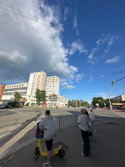 The image depicts an outdoor scene in Rubtsovo, Russia. The primary subjects are three people, two women and one man, interacting on a street. One woman is standing and looking away from the camera, wearing a patterned shirt and dark pants. Another woman, with blonde hair, is also standing, wearing a white patterned shirt and dark pants, and has a bag slung over her shoulder. The man is seated on a yellow electric scooter, wearing a grey hooded sweatshirt and black shorts, looking down at something on the scooter.
The setting appears to be a city street with a sidewalk and a road. In the background, several tall buildings line the street, some of which have visible signage. Green trees are also present, suggesting a relatively urban environment. The sky is a vibrant blue with scattered white clouds, indicating fair weather. Power lines are prominently visible, crisscrossing the sky.
The activity appears to be casual. The people are on a street, with one individual on a scooter, suggesting they might be commuting, sightseeing, or simply taking a break. The time of day is likely afternoon, given the bright sunlight and long shadows cast on the ground. There's no indication of a specific event or strong emotions.
Visible text includes what appears to be the number "00278458" on the yellow scooter. Other text is present on building signage and a street sign, but it is too small or blurry to be legible in detail. The overall impression is a snapshot of everyday life on a street in a Russian city.