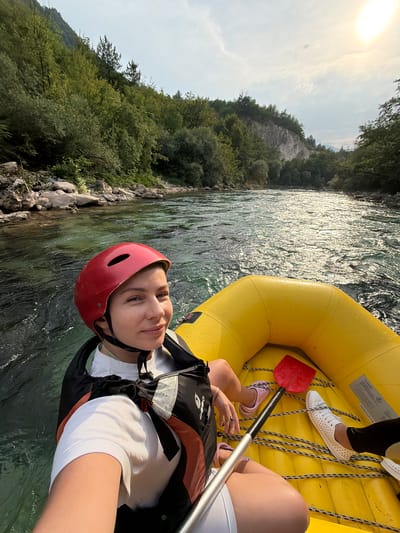 This media file shows a young woman in a yellow raft on a river. She is wearing a red helmet and an orange and black life vest over a white t-shirt. Her left hand is holding a paddle with a red blade. In the background, a rocky riverbank is covered with lush green trees and foliage. The sun is visible in the sky, suggesting it is daytime. The water is clear and appears to be flowing. Another person's legs and feet are visible in the raft, one wearing white perforated shoes and the other wearing pink slip-on shoes. This image was taken in Polje Bijela, Bosnia and Herzegovina. The overall mood appears adventurous and joyful.