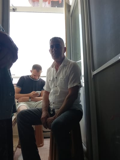 In Bahşılı Belediyesi, Turkey, a man with short, graying hair and visible wrinkles on his face sits on a stool, looking forward. He wears a white short-sleeved button-down shirt and dark pants. To his left, another man, with short dark hair and a beard, is seated and looking down at a white smartphone in his hands. He wears a dark t-shirt with white trim and light-colored shorts. A third person, mostly out of frame on the left, is dressed in dark clothing. The scene appears to be indoors, near a door or window with a screen, and the lighting suggests daytime.