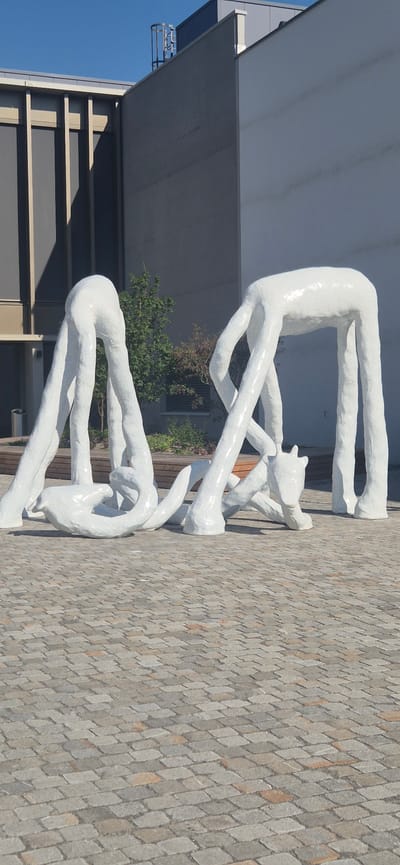 This image features a white, abstract sculpture composed of intertwined, elongated forms that resemble human limbs or bodies. The sculpture appears to be made of a smooth, possibly ceramic material, with a subtle texture visible upon close inspection. It is situated outdoors on a paved ground made of diamond-shaped interlocking stones in various shades of grey and brown.
In the background, there is a modern building with large, flat grey surfaces and a clear blue sky above. A section of the building has a projecting overhang, and there's a glimpse of some greenery, suggesting trees or bushes, between the sculpture and the building. The lighting indicates it's daytime, with shadows cast by the sculpture and the building.
The sculpture itself is the primary subject, depicting figures in a contorted, dynamic pose, almost as if wrestling or engaged in a fluid dance. The forms are smooth and flowing, creating a sense of movement and organic unity. There is no visible text in the image. The overall impression is one of artistic expression in an urban or semi-urban environment.