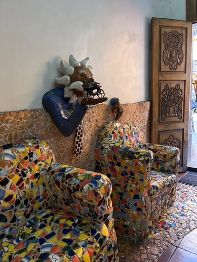 A mosaic-covered seating area fills the frame, featuring armchairs and adjacent wall sections adorned with multicolored tiles. A decorative wooden panel hangs above the mosaic. A sculpture is mounted on the wall, depicting a head with radiating sun-like features and a blue arm. The scene is visually rich with texture and artistic elements, possibly showcasing unique architectural or interior design.