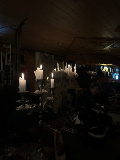 The image captures a dimly lit indoor scene, likely a restaurant or cozy gathering place in the evening or night. The primary light source comes from numerous candles arranged on tables and decorative structures, casting a warm, flickering glow across the room.
In the foreground and mid-ground, multiple candelabras hold pillar and tapered candles, many showing significant melt-down with wax cascading over their sides and holders. One prominent feature is a large, white, textured centerpiece, possibly an ice sculpture or a decorative element resembling snow or rock, studded with many small, upright candles. Wine glasses are visible on the tables, suggesting a dining or social setting.
Several people are seated at tables in the background and mid-ground, mostly silhouetted or partially visible due to the low light. One individual in the mid-ground, wearing a dark jacket, is more discernible, looking towards the right. A "Reusch" logo is visible on an item of clothing or accessory worn by one of the individuals.
The setting appears rustic, with wooden paneling on the walls and what looks like stone or brickwork in the background. The overall ambiance is intimate and atmospheric, created by the abundant candlelight. A sign with the French word "SORTIE" (Exit) is visible on a wooden column or doorframe, confirming a French-speaking location, consistent with Val-d'Isère, France. The scene depicts an event or evening meal, characterized by its warm, candlelit intimacy.