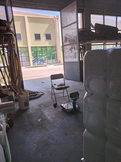 A wide shot captures the interior of a warehouse or industrial space in Döşemealtı, Turkey. Sunlight streams in from an open doorway and large windows, illuminating the concrete floor and the exterior building. The main subjects are various objects within the space: a stack of large, white, plastic-wrapped bales or pallets dominate the right side of the frame, their smooth, shiny surfaces reflecting the light. To the left, a collection of hoses, possibly for industrial use, hang vertically from a rack. In the foreground, two white buckets are visible, one containing green material and another with a red lid, and coiled hoses rest on the floor. A small, foldable metal chair with a worn seat is placed near a metal platform scale. The scale itself appears to be an older model with a digital display.
Beyond the immediate interior, the scene opens to an exterior view. A pale yellow building with rectangular windows and a light-colored trim is visible through the open doorway. Large windows on this exterior building are decorated with a mural depicting green leaves and a blue butterfly, suggesting a focus on nature or perhaps an environmentally conscious business. The exterior ground appears to be paved.
The lighting suggests it is daytime, likely morning or afternoon given the strong sunlight. The overall impression is one of a functional, working space, possibly involved in storage or distribution, with elements of nature incorporated into its exterior design. No people are visible in the image. There is some faded text on the bucket, but it is not legible.