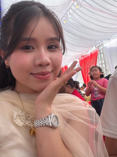 A young woman is captured in a close-up selfie, angled to show her upper body and the surrounding environment, likely in Tay Son Commune, Vietnam. She has dark hair, styled down, and dark eyes, with a subtle smile and pinkish-red lipstick. She wears a light-colored, possibly cream or beige, sheer fabric dress or top, which appears to be layered or ruffled. On her left wrist, she sports a silver-toned wristwatch with a detailed, possibly jeweled, bezel and a metal link band. Her right hand is raised, making a peace sign towards the camera.
In the background, a young girl with dark hair tied in pigtails is visible, looking directly towards the camera with a slight pout or curious expression. She is wearing a pink t-shirt with a colorful graphic print. Other blurred figures of people suggest a gathering or event.
The setting appears to be under a large canopy or tent, with extensive white draped fabric forming the ceiling. Sections of red and white striped fabric are visible in the background, contributing to the festive or event atmosphere. Small, round light fixtures, some yellow and some white, are strung along the canopy structure. The bright, even lighting indicates it is likely daytime. The overall scene suggests a casual event or celebration taking place under a temporary shelter. There is no visible text in the image.
