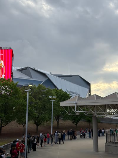The image captures the Mercedes-Benz Stadium in Atlanta, Georgia, U.S.A. A queue of people can be seen lining up in front of the stadium on what appears to be a concrete walkway. Overhead, a covered walkway structure crosses above the queuing crowd. Tall trees with green foliage flank the walkway. A large digital display board with red light and graphics is visible to the left of the stadium. The sky is overcast with dense gray clouds.