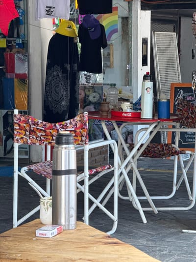 An outdoor market stand displays various merchandise. Clothes, including t-shirts and patterned pants, hang near a reflective glass surface. A rack showcases items, with a stainless steel thermos and a pack of "BOXER" cigarettes visible. Coca-Cola branding is present. A chalkboard menu lists "VINOS," "GABEOSA," and "CERVEZA." A rainbow sticker appears in the background. Text "MODAS," "REDDELDO," "POTRERO," "FORES," "AZÚCAR," and "SANDW" are identifiable, suggesting the market is in a Spanish-speaking area.