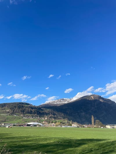 The image depicts a view in Sterzing-Vipiteno, Italy. The foreground consists of a bright green grass field. Behind the field, there's a town with buildings, including a prominent arched structure. A mountain range rises behind the town, partly covered with patches of snow. The sky is a clear blue, dotted with scattered clouds. There appears to be a contrail in the sky.